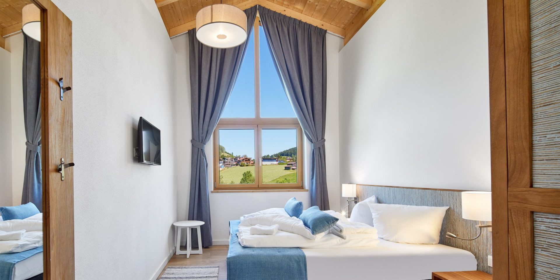 Residenz Drachenstein Niederau Wildschonau Oostenrijk wintersport skivakantie luxe slaapkamer D8A 2-persoonsbed blauwe kussens krukje lamp houten balken hoge ramen nachtlampjes