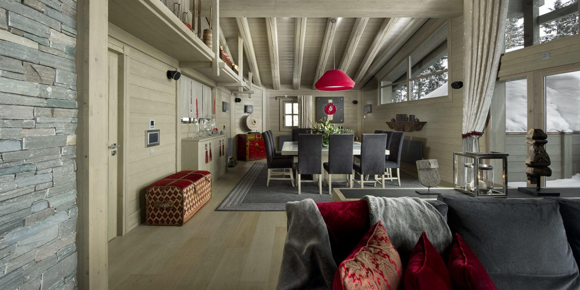 Le K2 Palace Suite-Chalet Baltoro Courchevel Les 3 Vallees Frankrijk wintersport skivakantie luxe eetkamer grote vierkante eettafel stoelen rode hanglamp rode kist grijze bank rode kussens ramen houten balken bomen sneeuw