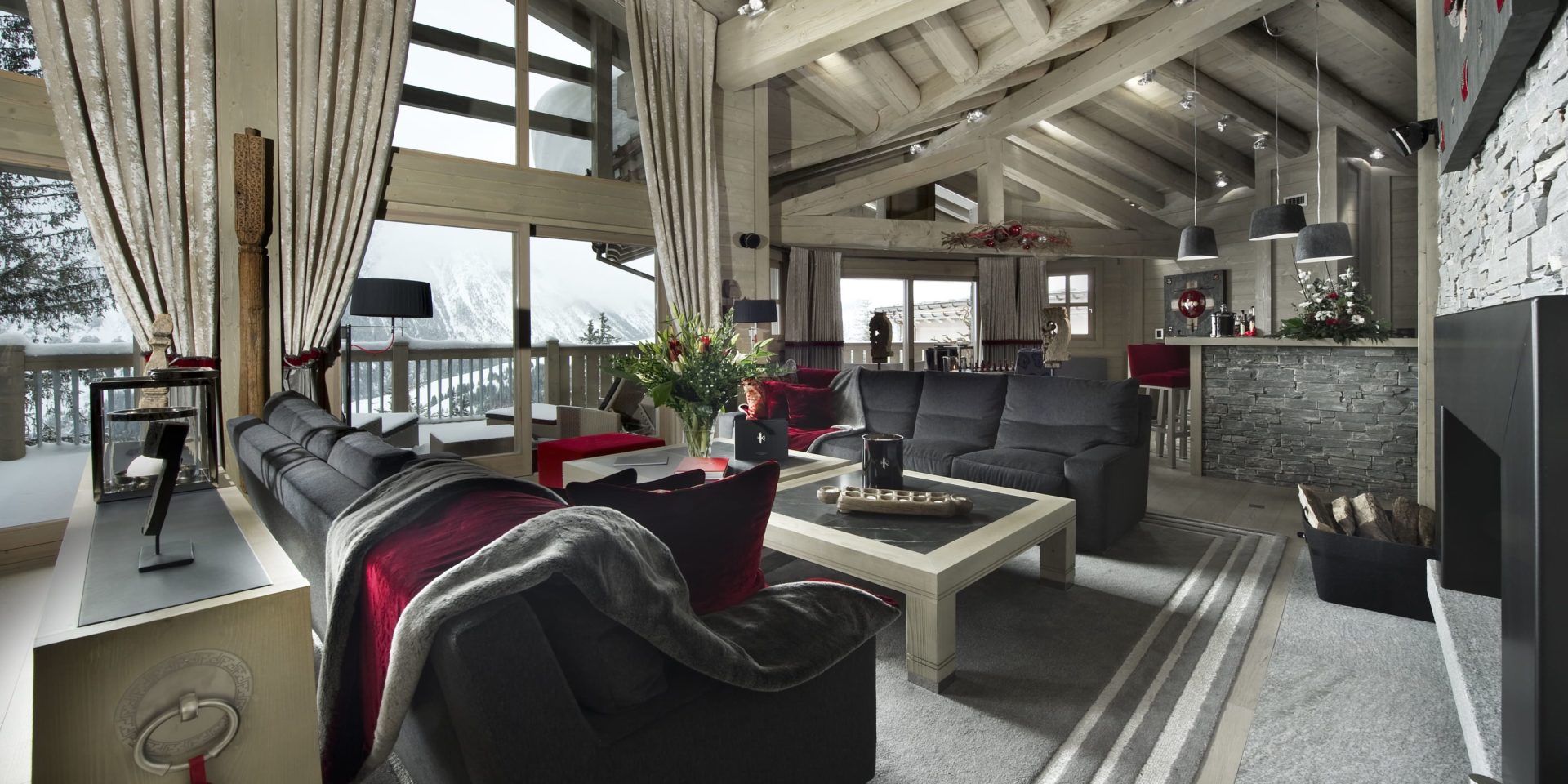 Le K2 Palace Suite-Chalet Baltoro Courchevel Les 3 Vallees Frankrijk wintersport skivakantie luxe living grijze banken rood kleed kussens salontafel bloemen bar rode barkrukken hanglampen hout open haard ramen houten balken bomen sneeuw