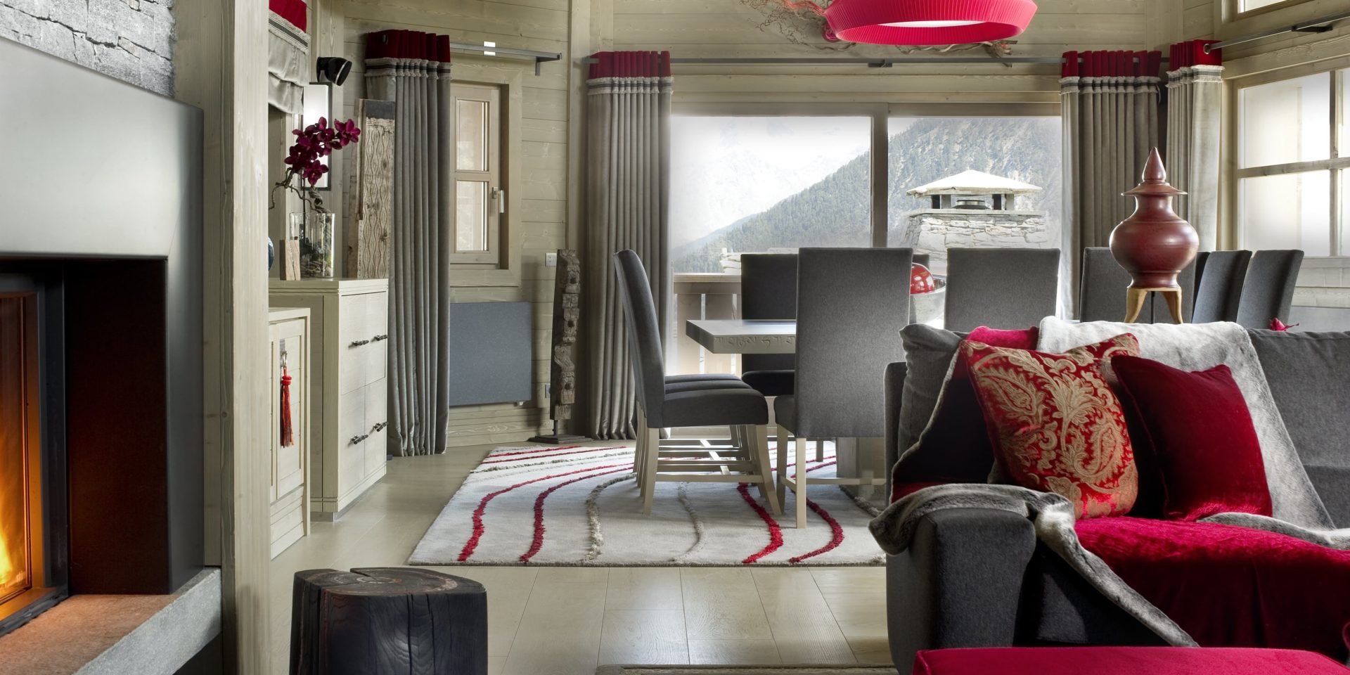 Le K2 Palace Suite-Chalet Muztagh Courchevel Les 3 Vallees Frankrijk wintersport skivakantie luxe eetkamer grote vierkante eettafel stoelen rode hanglamp grijze bank rode kussens open haard vloerkleed raam hout bergen sneeuw