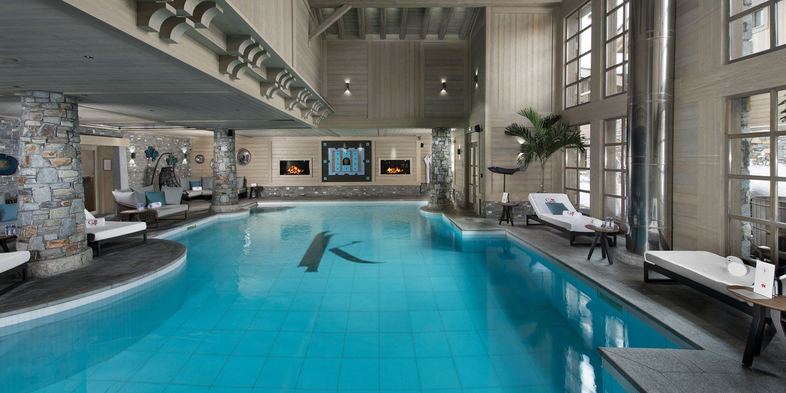 Hotel Le K2 Altitude Courchevel Les 3 Vallees Frankrijk wintersport skivakantie luxe zwembad hoekbank loungestoelen blauwe kussens hangende stoel stenen pilaren houten plafond open haard hoge ramen zwemmen ontspannen relaxen genieten