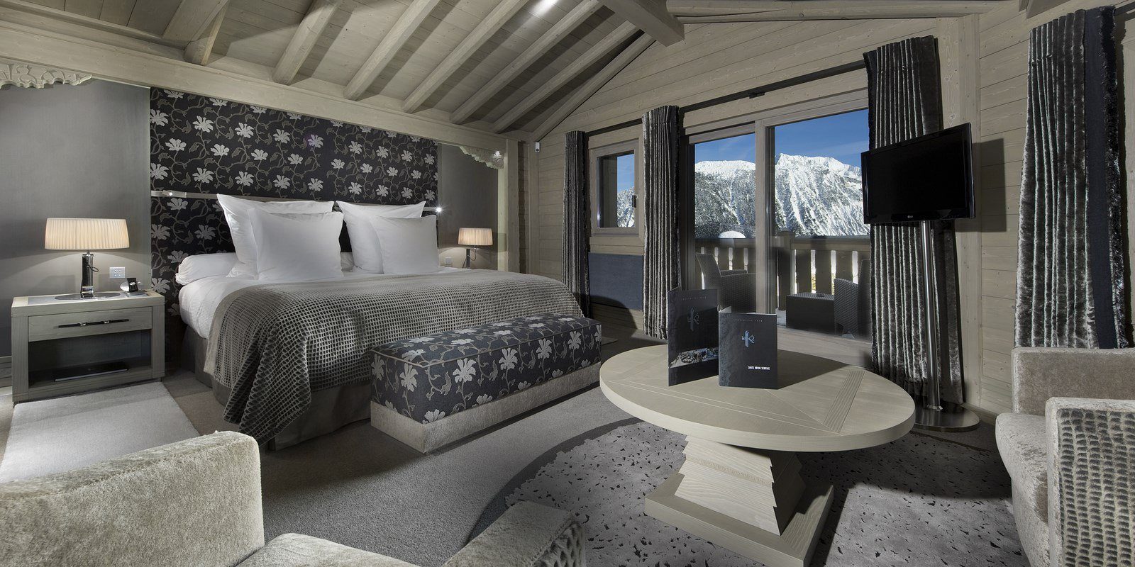 Hotel Le K2 Palace Courchevel Les 3 Vallees Frankrijk wintersport skivakantie luxe slaapkamer 2-persoonsbed zwart wit bloemenmotief hocker fauteuils salontafel houten balken houten plafond balkon besneeuwde bergen sneeuw