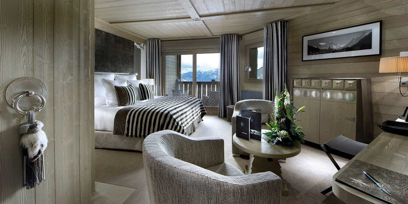 Hotel Le K2 Palace Courchevel Les 3 Vallees Frankrijk wintersport skivakantie luxe Suite 2-persoonsbed fauteuils salontafel bloemstuk schilderij bureau kruk houten plafond balkon stoelen besneeuwde bergen sneeuw