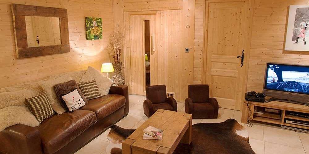 Chalet Belle Vache Chatel Les Portes du Soleil Frankrijk wintersport skivakantie luxe living bruine leren bank kinder fauteuils salontafel boeken vloerkleed TV spiegel houten wanden schilderij