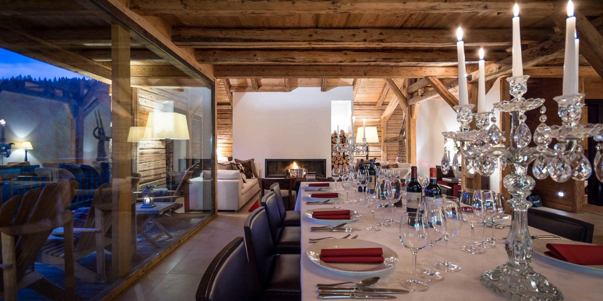Chalet Ferme de Moudon Les Gets Les Portes du Soleil Frankrijk wintersport skivakantie luxe eetkamer gedekte tafel kandelaar kaarsen wijn wijnglazen open haard witte bank houten balken houten wanden raam balkon ligstoelen bomen