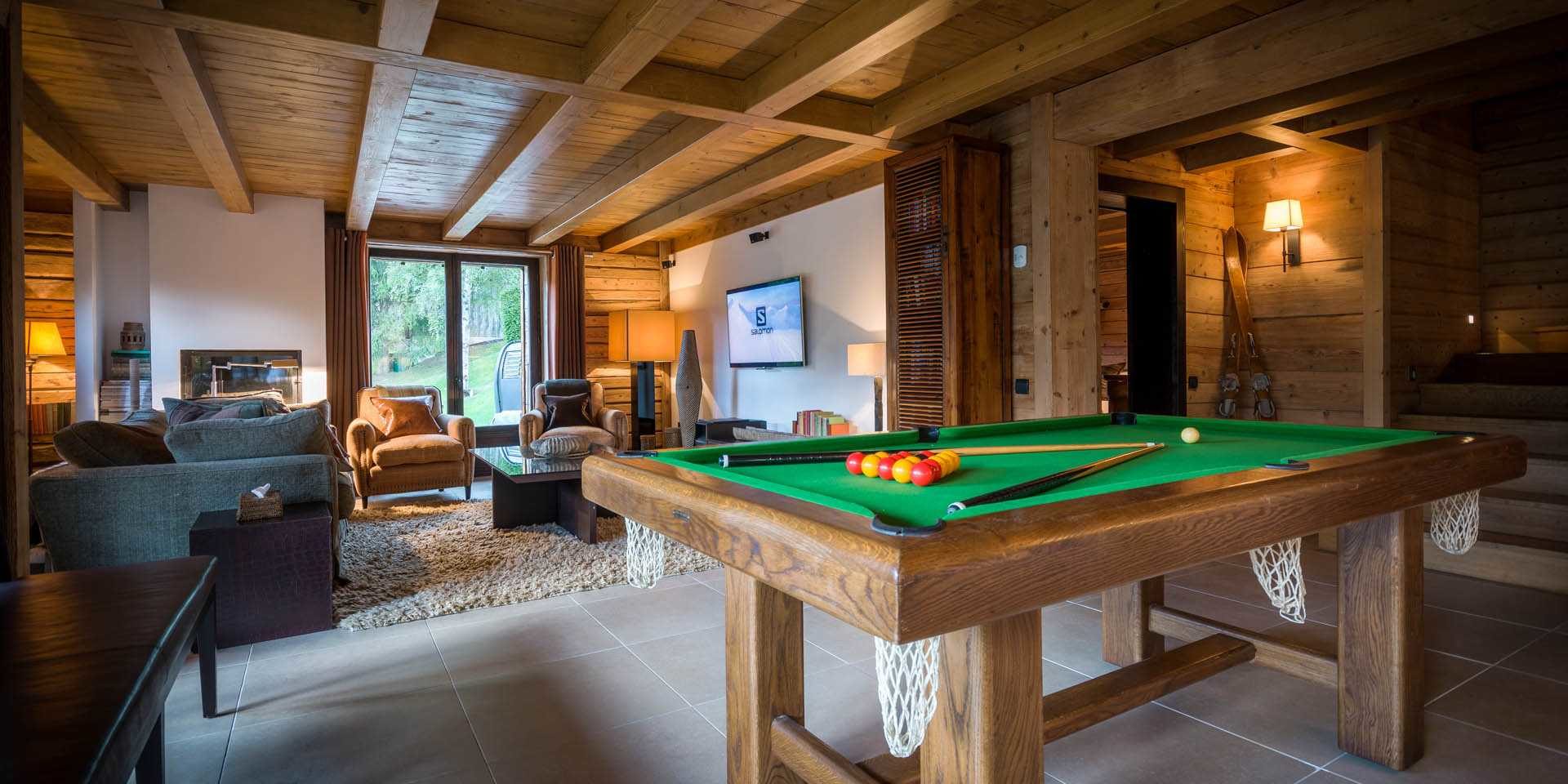 Chalet Ferme de Moudon Les Gets Les Portes du Soleil Frankrijk wintersport skivakantie luxe zitkamer pooltafel wanlamp zithoek banken fauteuil TV salontafel vloerkleed raam bomen houten balken