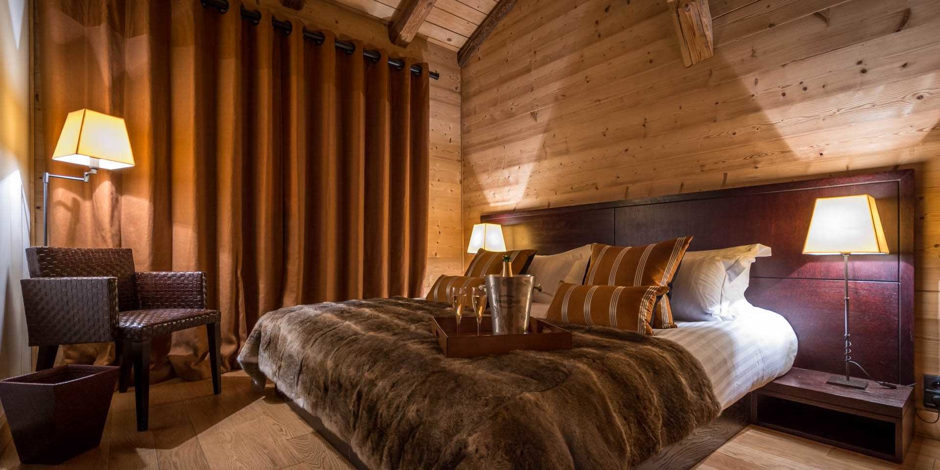 Chalet Ferme de Moudon Les Gets Les Portes du Soleil Frankrijk wintersport skivakantie luxe slaapkamer 2-persoonsbed grijze sprei Champagne glazen fauteuil nachtlampjes gordijnen houten wanden houten balken