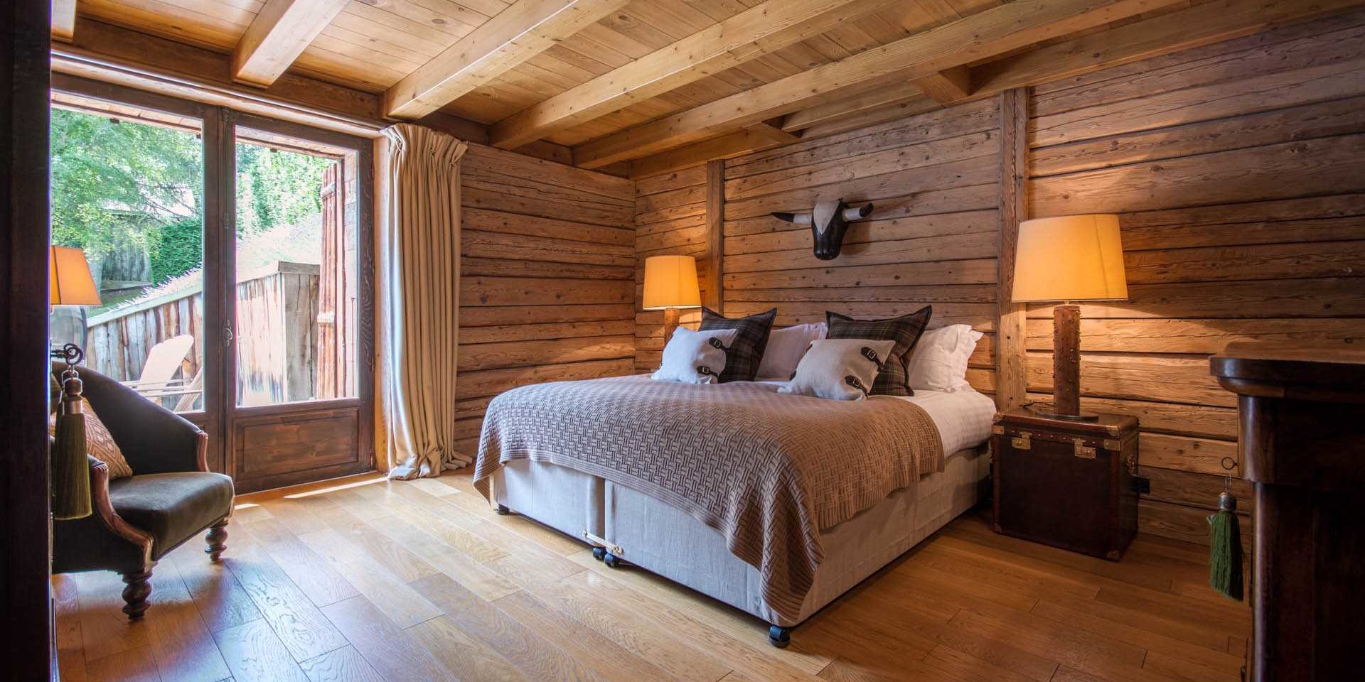 Chalet Ferme de Moudon Les Gets Les Portes du Soleil Frankrijk wintersport skivakantie luxe slaapkamer 2-persoonsbed bruine sprei fauteuil stierenkop nachtlampjes kist houten wanden houten balken terras ligstoelen bomen