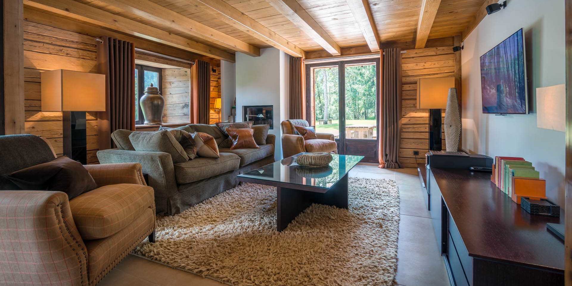 Chalet Ferme de Moudon Les Gets Les Portes du Soleil Frankrijk wintersport skivakantie luxe zitkamer grijze bank kussens fauteuils salontafel TV vloerkleed vaas lampen houten wanden houten balken raam bomen