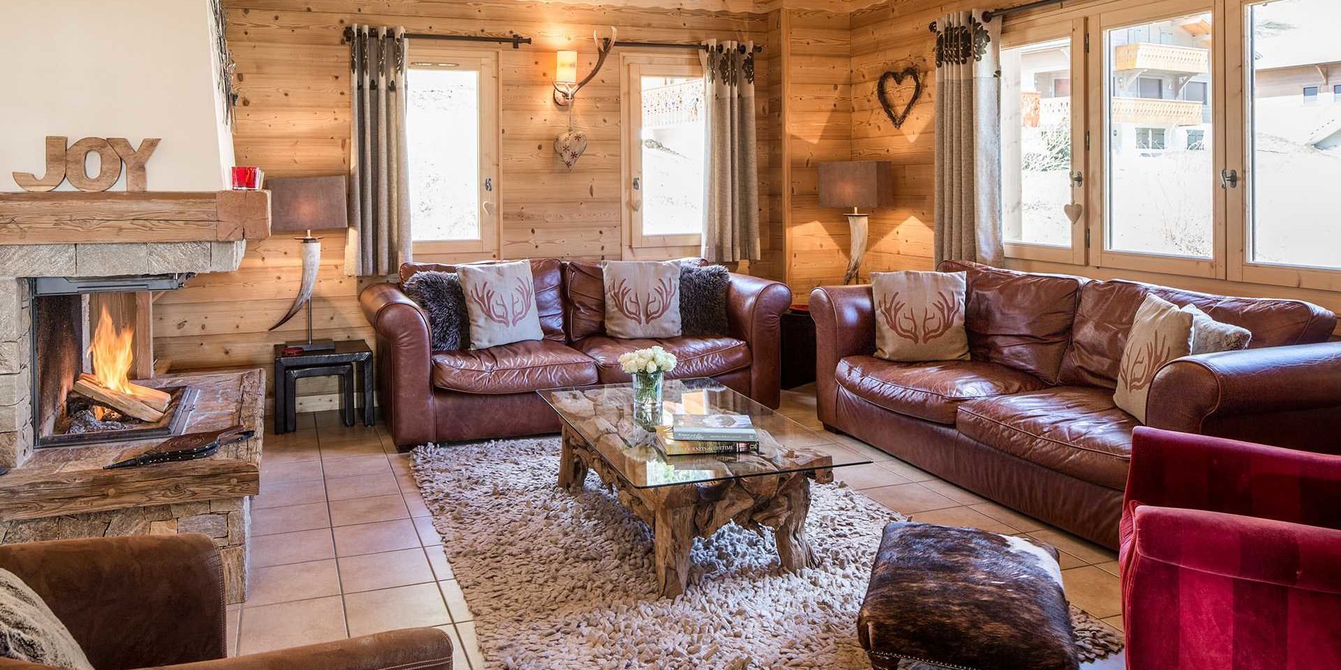 Chalet Grand Coeur Chatel Les Portes du Soleil Frankrijk wintersport skivakantie luxe living bruine leren banken kussens gewei rode bruine fauteuils hocker open haard salontafel bloemen boeken hart vloerkleed gewei JOY ramen houten wanden