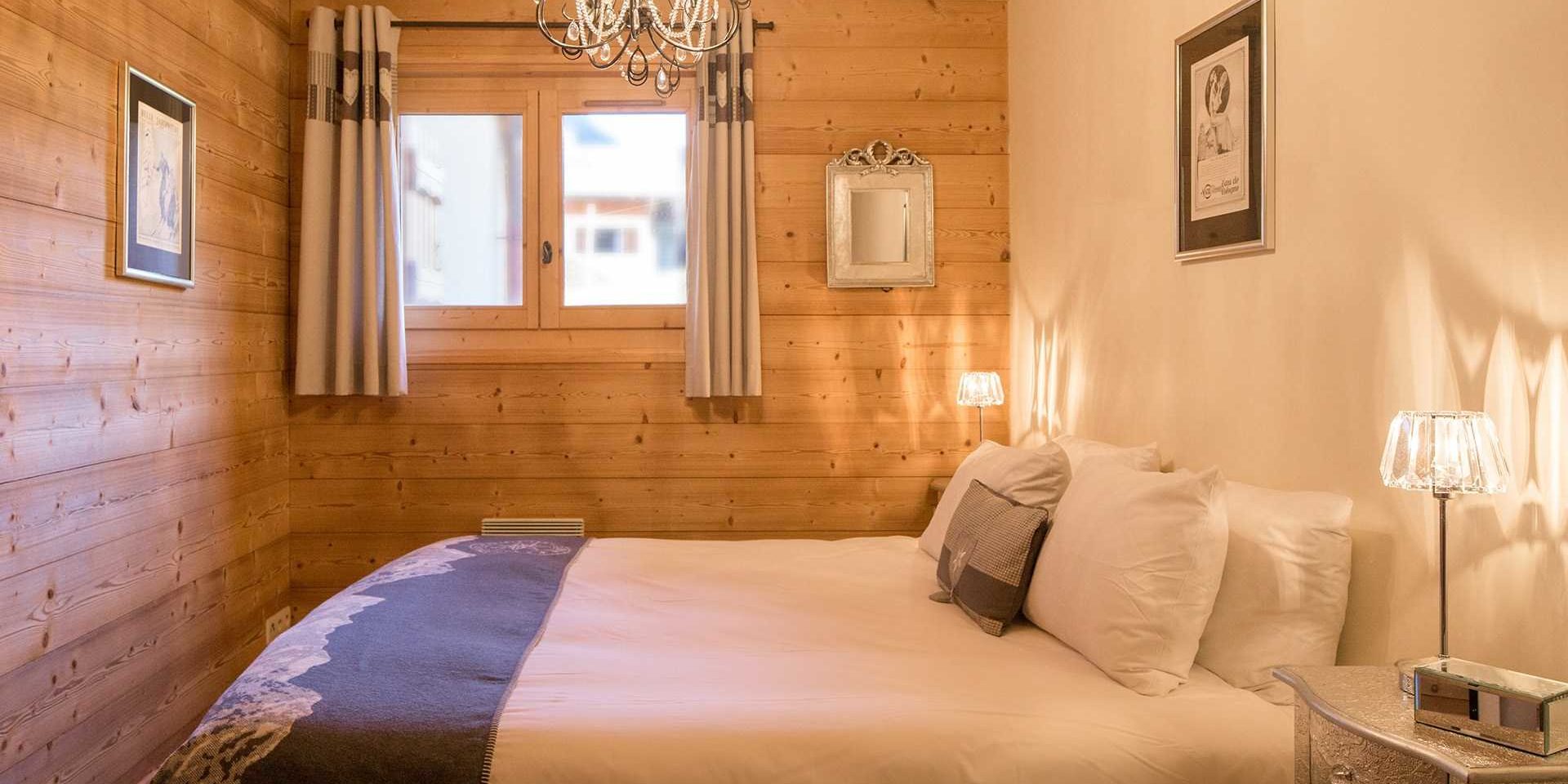 Chalet Grand Coeur Chatel Les Portes du Soleil Frankrijk wintersport skivakantie luxe slaapkamer 2-persoonsbed sprei witte grijze kussens nachtlampjes schilderijen houten wanden houten balken raam kroonluchter spiegel