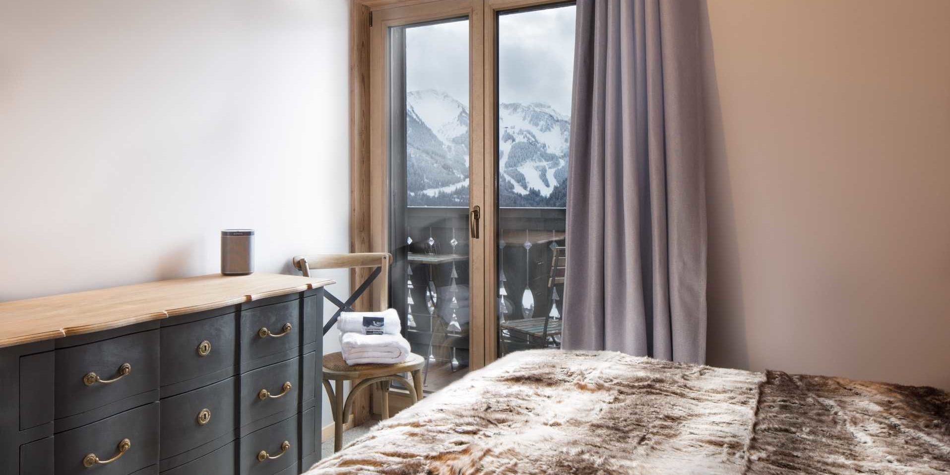 Chalet Les Clarines Chatel Les Portes du Soleil Frankrijk wintersport skivakantie luxe slaapkamer 2-persoonsbed dressoir stoel handdoeken paarse gordijnen raam balkon stoeltjes besneeuwde bergen sneeuw