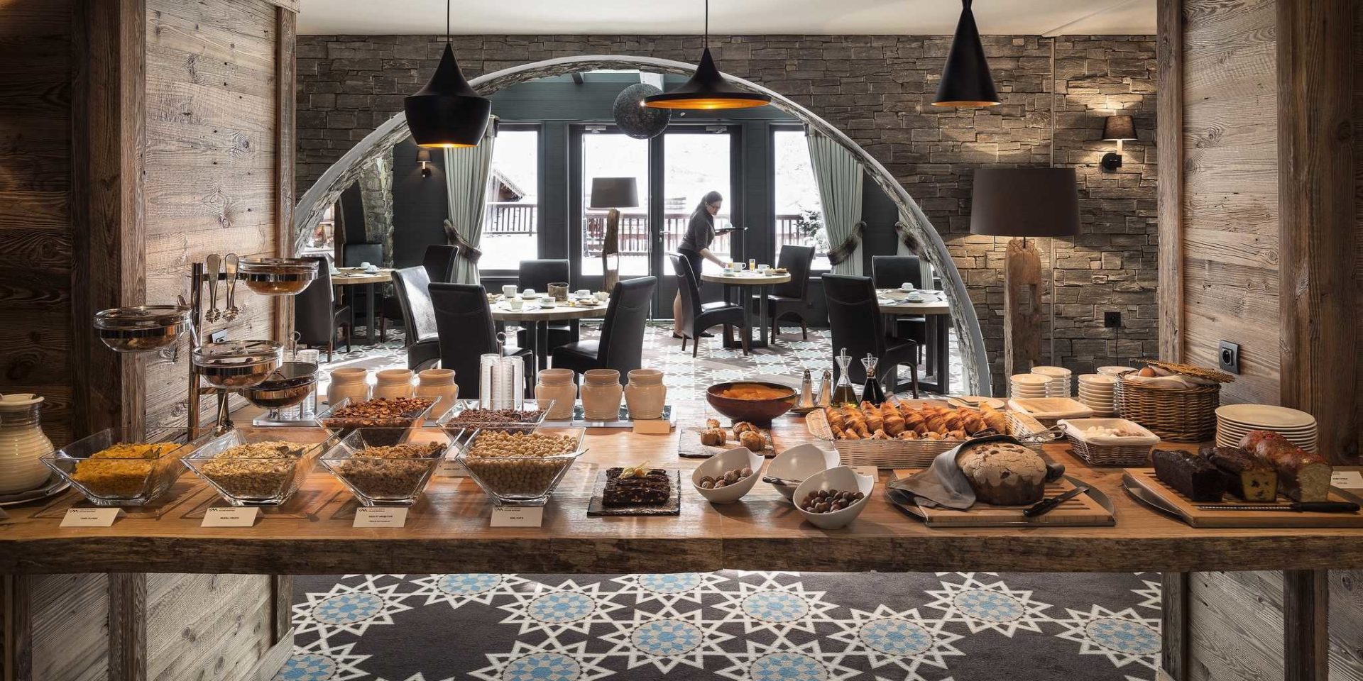 Hotel Village Montana Tignes Tignes-Val d'Isere Frankrijk wintersport skivakantie luxe restaurant Les Chanterelles ontbijt croissants muesli brood jam hanglampen half ronde doorgang patroon vloerbedekking gedekte tafels serveerster