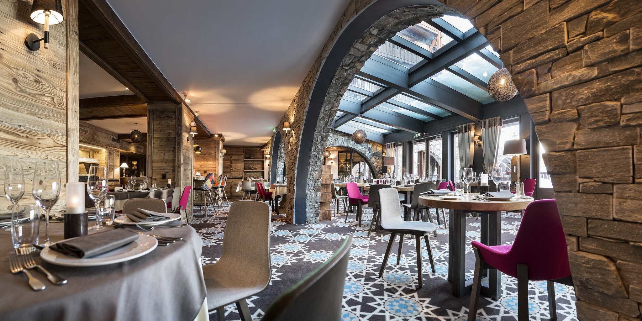 Hotel Village Montana Tignes Tignes-Val d'Isere Frankrijk wintersport skivakantie luxe restaurant Les Chanterelles gedekte tafels wijnglazen patroon vloerbedekking half ronde bogen roze grijze stoelen gekleurde stoelen hanglampen wandlampen