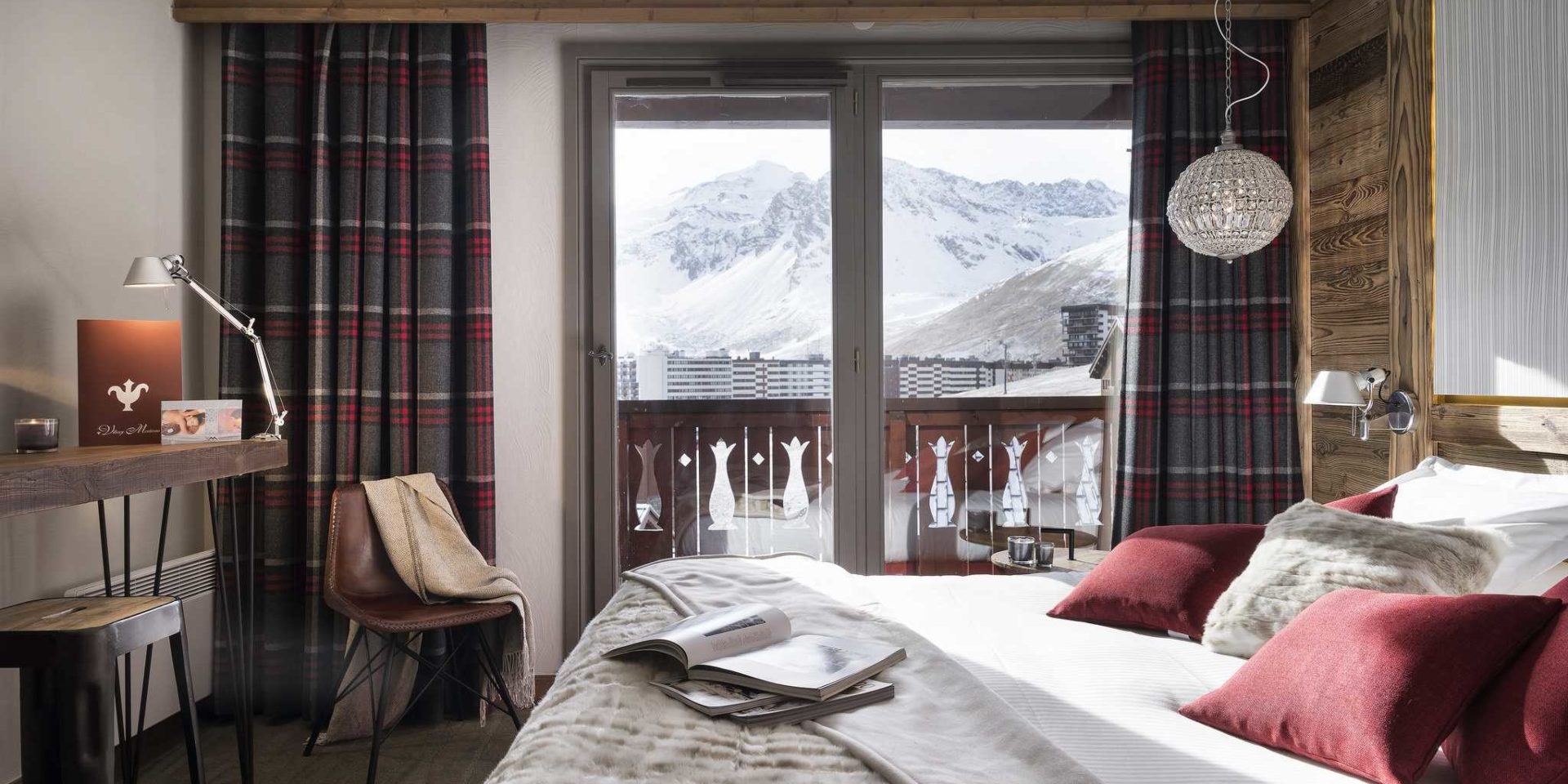 Hotel Village Montana Tignes Tignes-Val d'Isere Frankrijk wintersport skivakantie luxe slaapkamer 2-persoonsbed rode kussens sprei handlamp bruine stoel geblokte gordijnen balkon besneeuwde bergen
