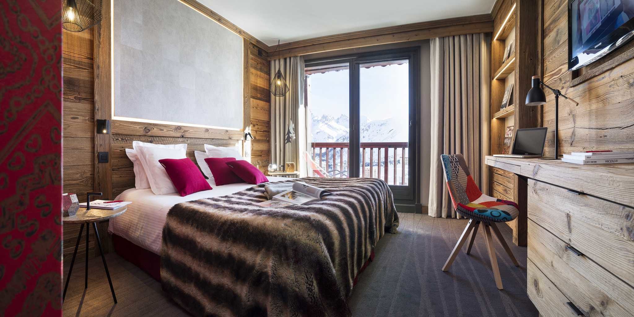 Hotel Village Montana Tignes Tignes-Val d'Isere Frankrijk wintersport skivakantie luxe slaapkamer 2-persoonsbed roze kussens sprei gekleurde stoel bureau TV hanglampen raam balkon besneeuwde bergen