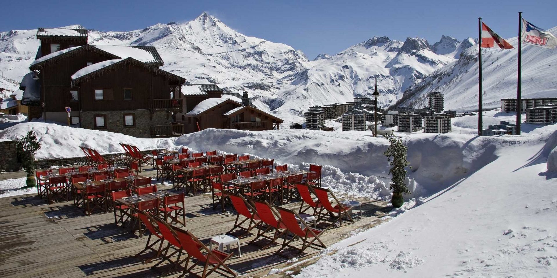 Hotel Village Montana Tignes Tignes-Val d'Isere Frankrijk wintersport skivakantie luxe restaurant La Chaumiere terras rode ligstoelen tafels rode stoelen plant vlaggen dorp Tignes besneeuwde bergen sneeuw