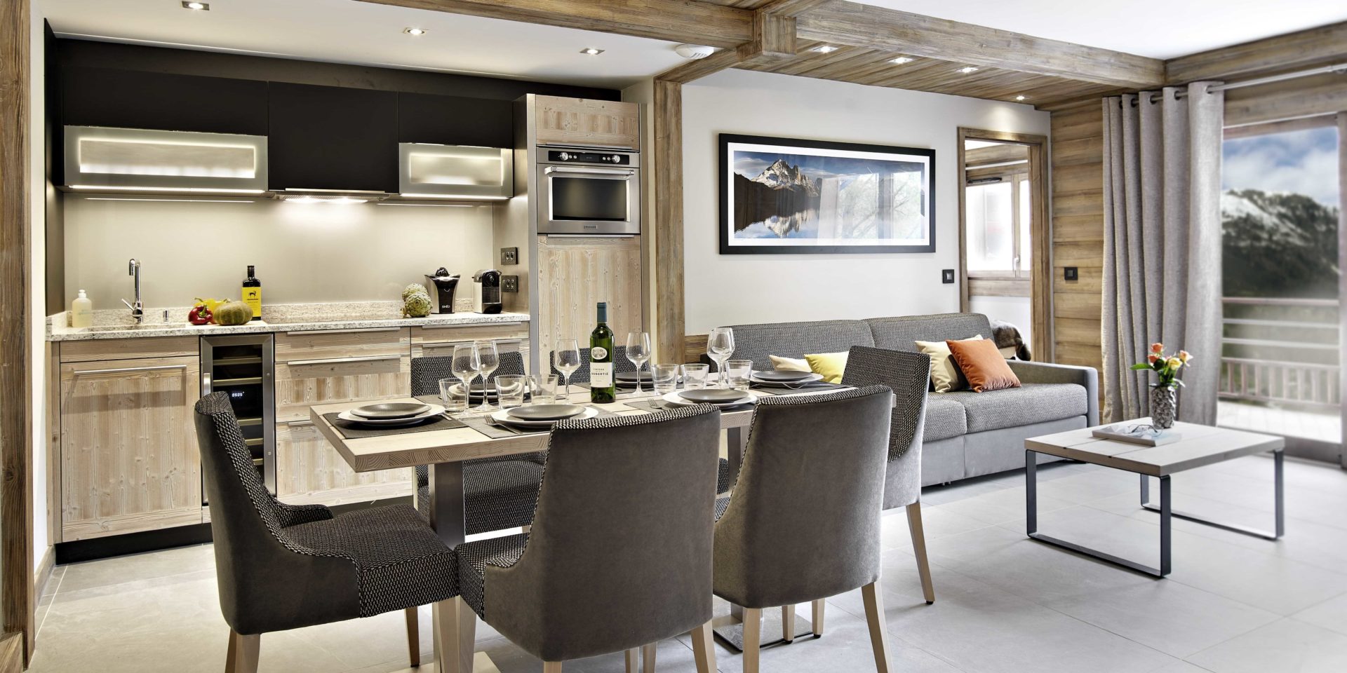 MGM Residence Les Chalets Elena Les Houches Chamonix Mont Blanc Frankrijk wintersport skivakantie luxe living gedekte tafel wijn wijnglazen stoelen bruine bank schilderij salontafel bloemen keuken raam balkon besneeuwde bergen