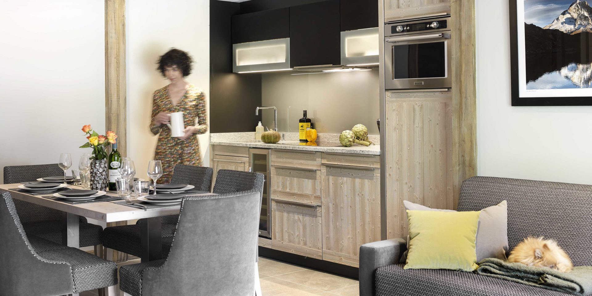MGM Residence Les Chalets Elena Les Houches Chamonix Mont Blanc Frankrijk wintersport skivakantie luxe gedekte tafel wijn rozen grijze bank geel kussen kleed schilderij keuken dame hond