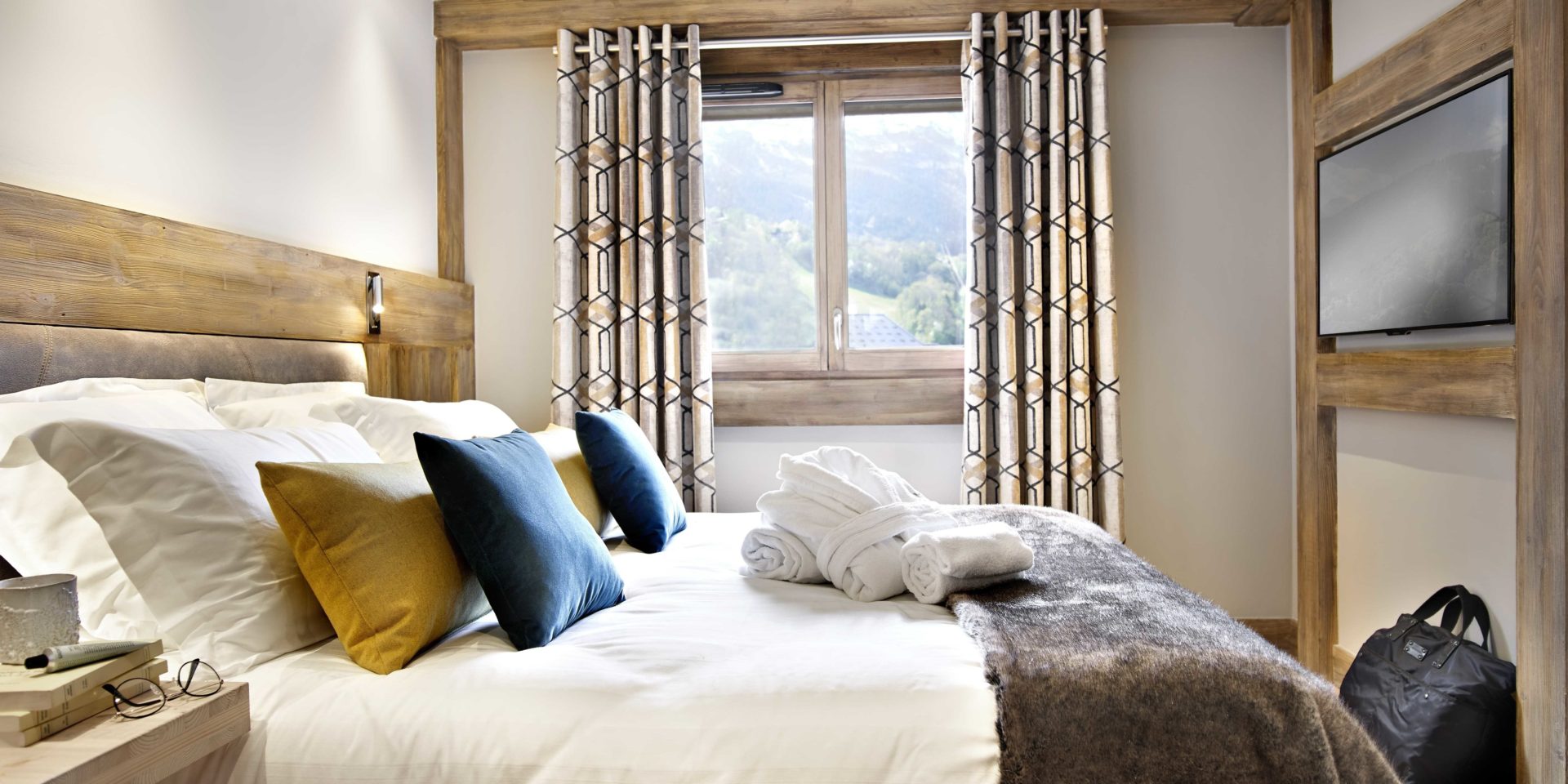 MGM Residence Les Chalets Elena Les Houches Chamonix Mont Blanc Frankrijk wintersport skivakantie luxe slaapkamer 2-persoonsbed gele blauwe kussens badjas handdoeken TV tas raam bomen bergen