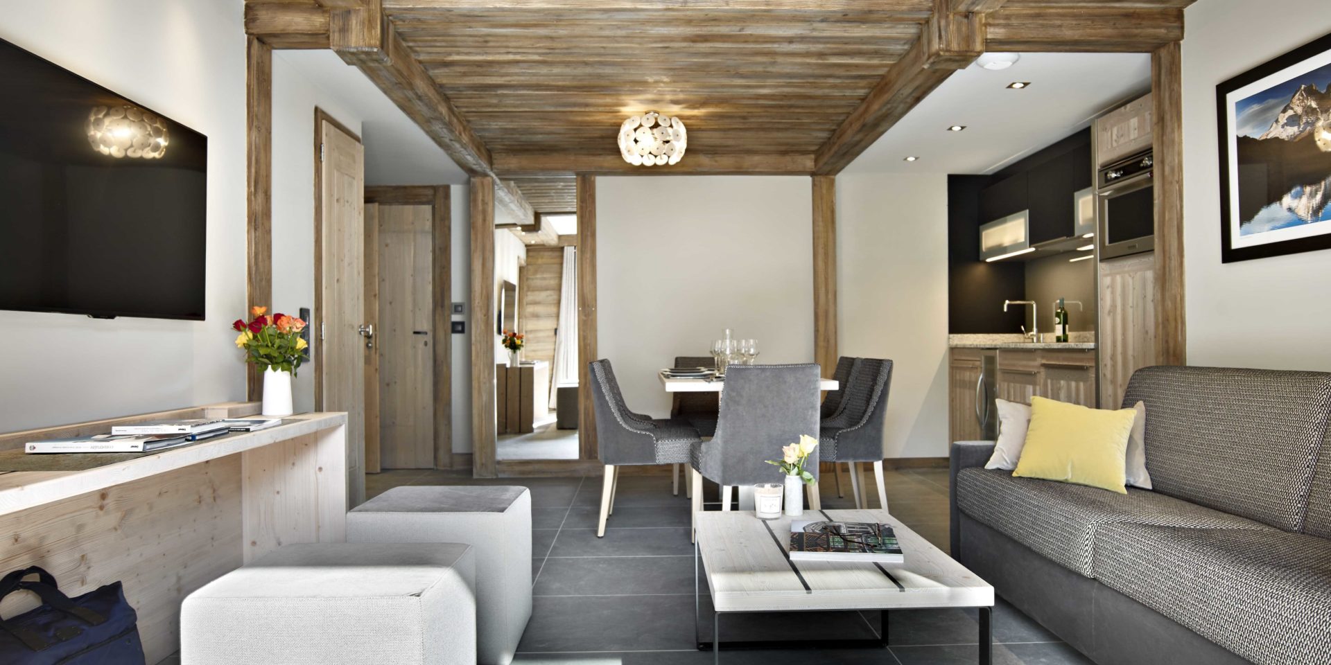 MGM Residence Les Chalets Elena Les Houches Chamonix Mont Blanc Frankrijk wintersport skivakantie luxe living grijze bank salontafel rozen hockers TV gedekte tafel wijnglazen stoelen hanglamp keuken tas houten plafond schilderij