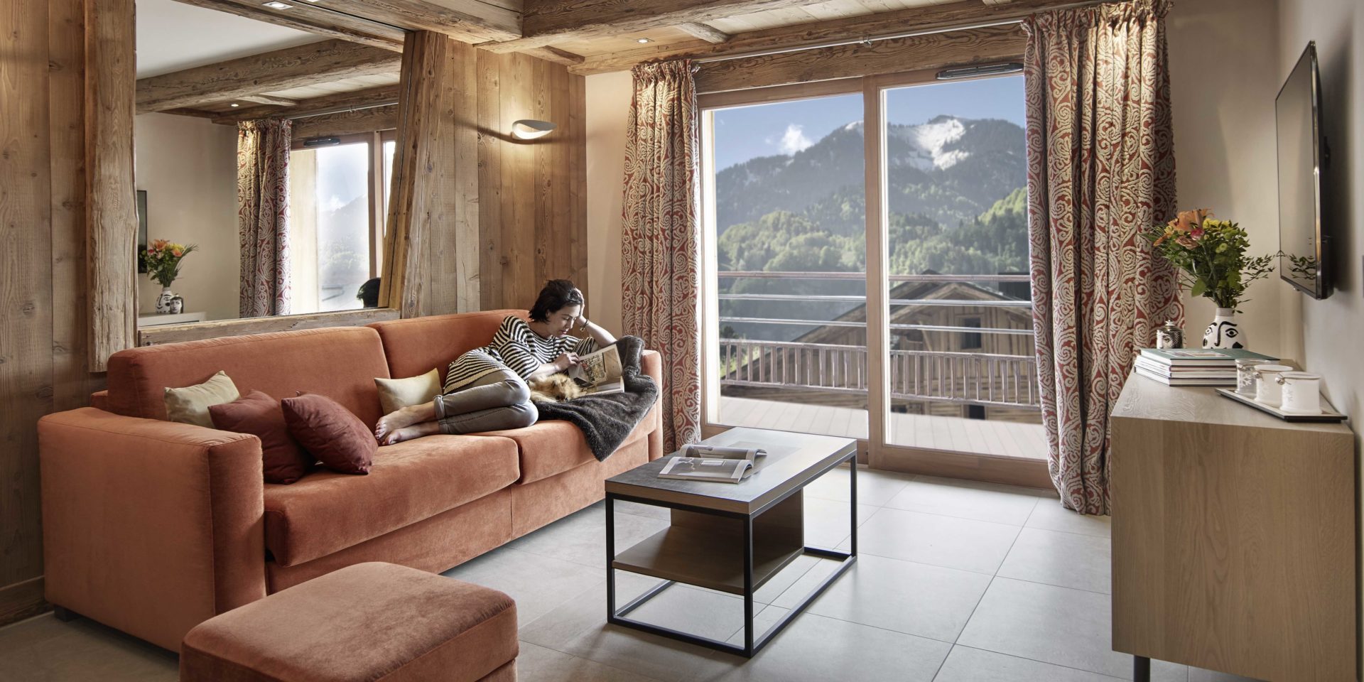 MGM Residence Alexane Samoens Le Grand Massif Frankrijk wintersport skivakantie luxe living rode bank hocker salontafel grote spiegel boeken TV bloemen dressoir raam balkon bergen bomen
