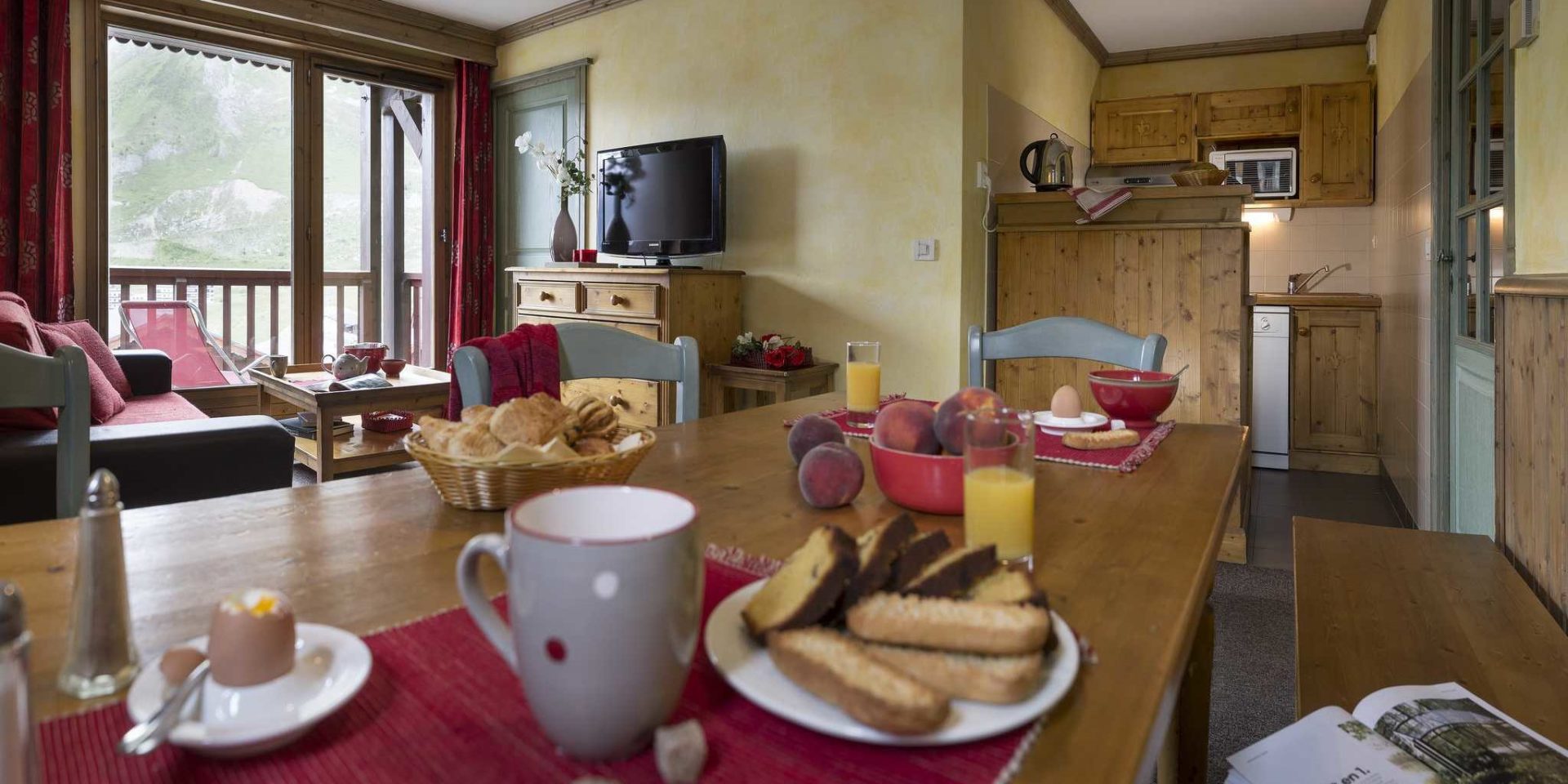 Residences Village Montana Tignes Tignes-Val d'Isere Frankrijk wintersport skivakantie luxe living 3K6 gedekte tafel ontbijt broodjes perziken eieren houten stoelen bankje rode bank salontafel thee TV vaas bloem balkon rode ligstoel keuken
