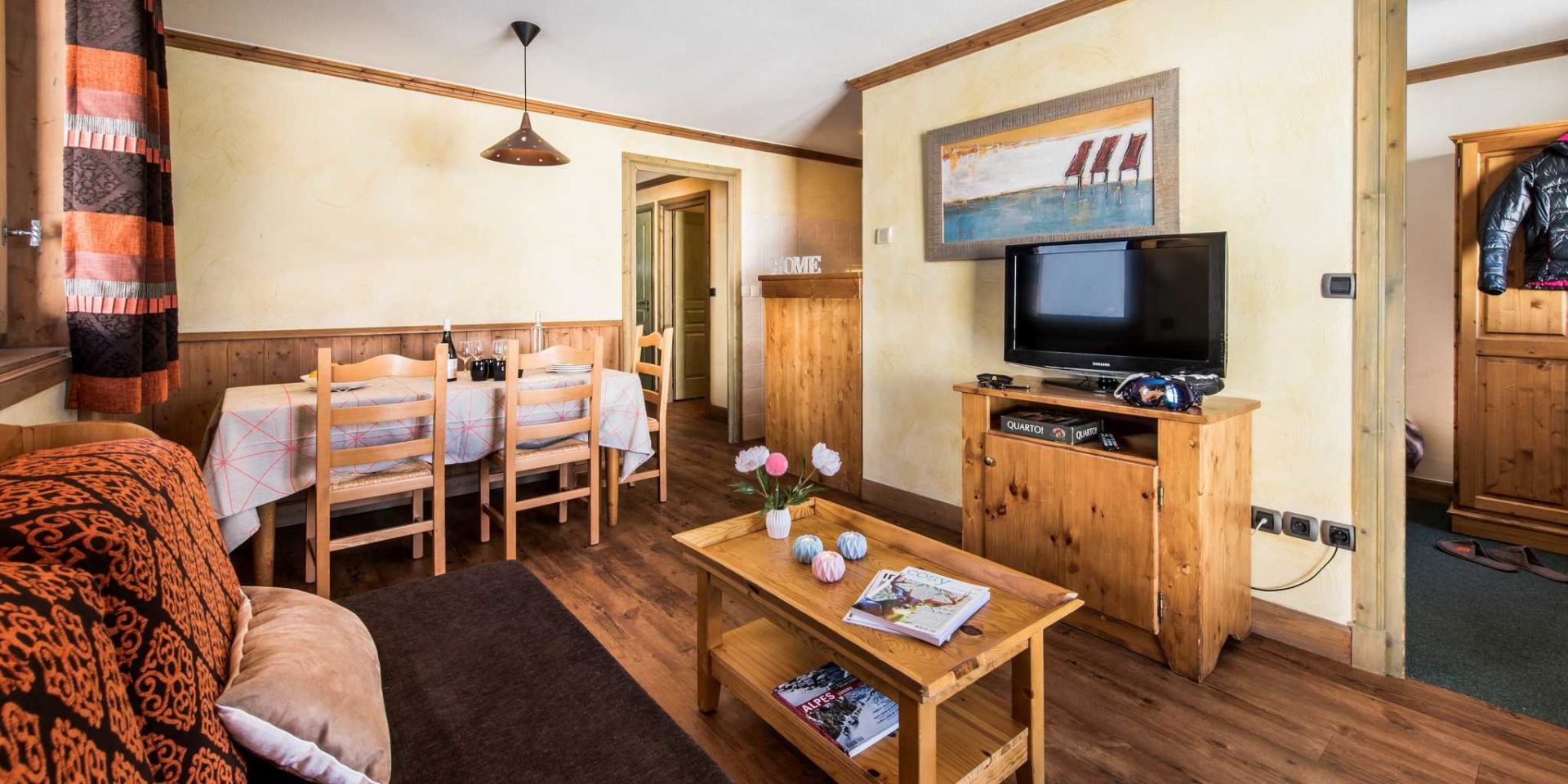Residences Village Montana Tignes Tignes-Val d'Isere Frankrijk wintersport skivakantie luxe living 6K10 bank oranje kussens salontafel kaarsen boeken bloemen TV schilderij eettafel wijn hanglamp houten stoelen keuken HOME slippers jas