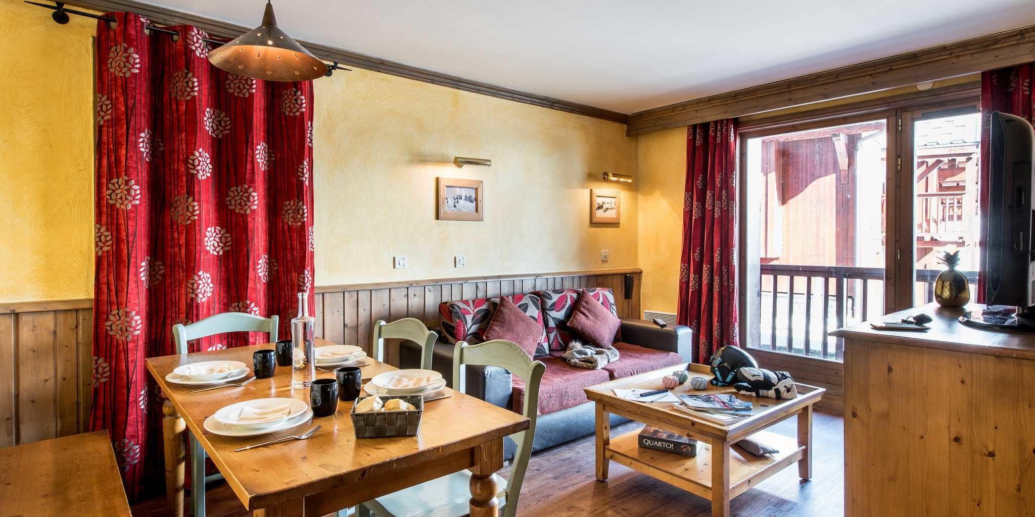 Residences Village Montana Tignes Tignes-Val d'Isere Frankrijk wintersport skivakantie luxe living 3K6 gedekte tafel karaf bank kussens salontafel boek TV rode gordijnen schilderijen balkon