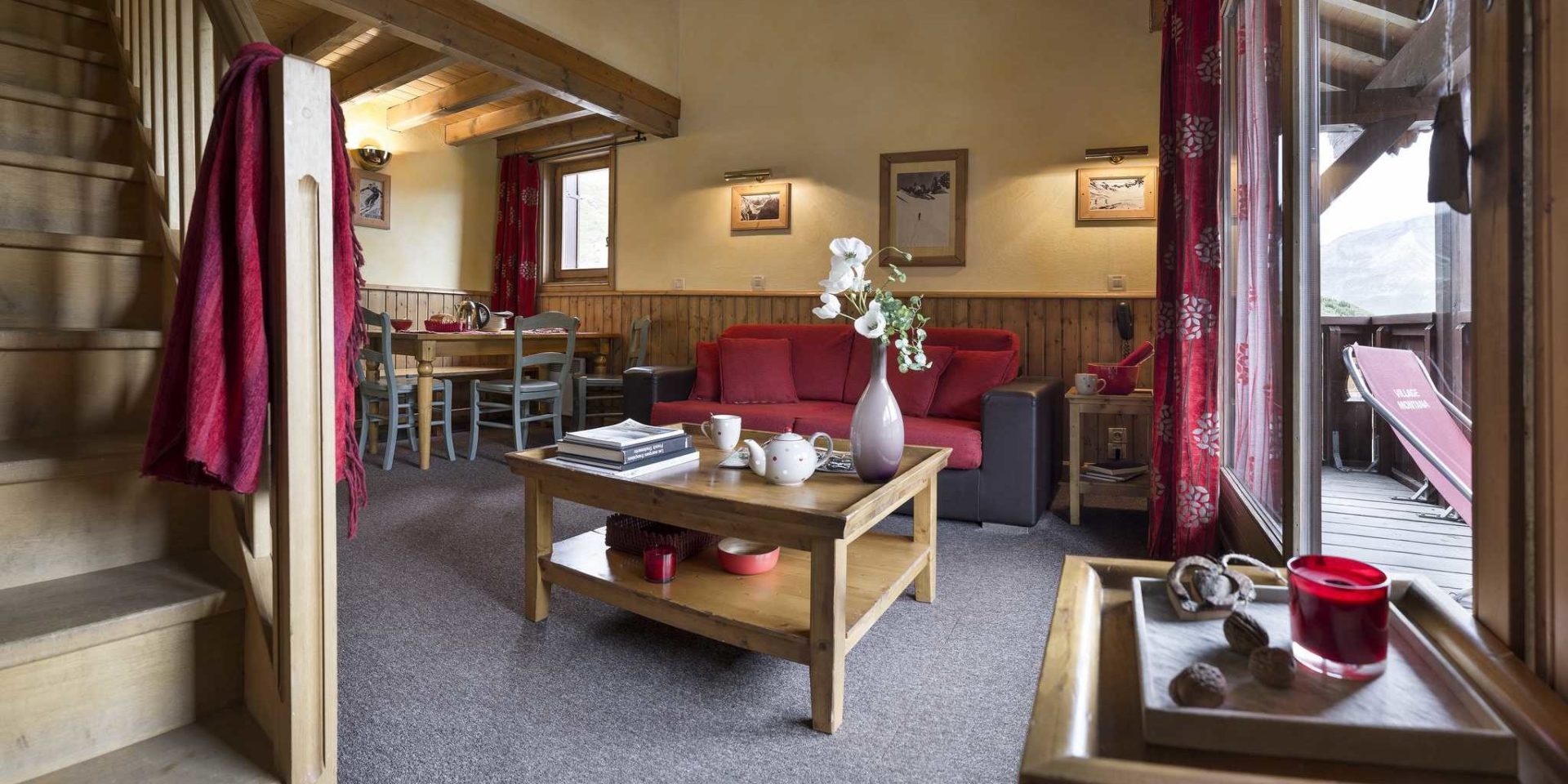 Residences Village Montana Tignes Tignes-Val d'Isere Frankrijk wintersport skivakantie luxe living 3K6 Cabine rode bank salontafel vaas bloemen kaars theepot boeken eettafel houten stoelen schilderijen houten trap houten balken balkon rode ligstoel bergen