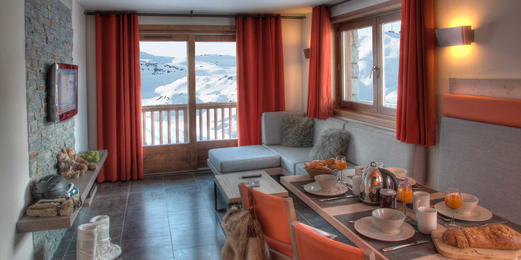 Residence Montana Plein Sud Val Thorens Les 3 Vallees Frankrijk wintersport skivakantie luxe living 3K4 gedekte tafel ontbijt broodjes oranje stoelen grijze bank kussens salontafel TV rode gordijnen balkon uitzicht besneeuwde bergen sneeuw