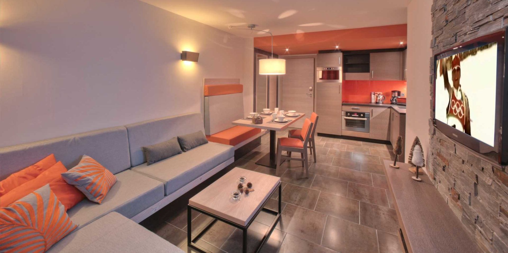 Residence Montana Plein Sud Val Thorens Les 3 Vallees Frankrijk wintersport skivakantie luxe living 3K4 grijze hoekbank oranje kussens salontafel TV gedekte eettafel oranje stoelen hanglamp keuken oranje muur dennenbomen
