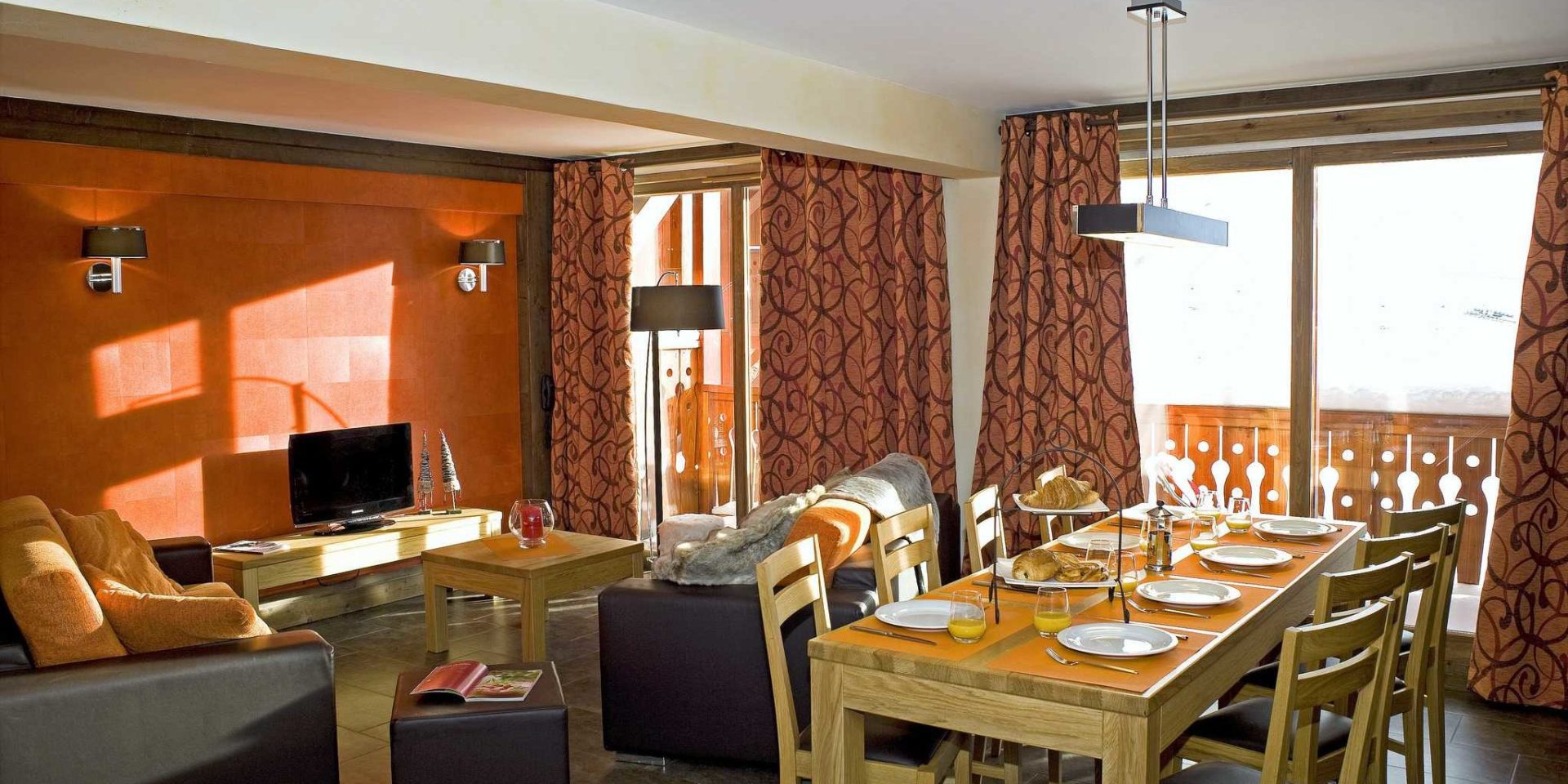 Residences Village Montana Val Thorens Les 3 Vallees Frankrijk wintersport skivakantie luxe living 5K8 gedekte tafel croissants broodjes houten stoelen bruine leren banken oranje kussens hocker salontafel kaars TV oranje muur balkon