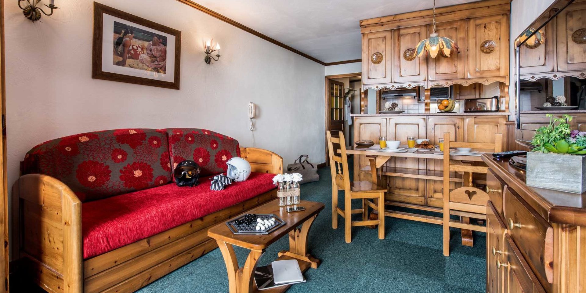 Residences Village Montana Val Thorens Les 3 Vallees Frankrijk wintersport skivakantie luxe living 2K3 bank rode kussens skihelmen handschoenen salontafel gezelschapsspel gedekte tafel houten stoelen hanglamp houten kast vers fruit schilderij