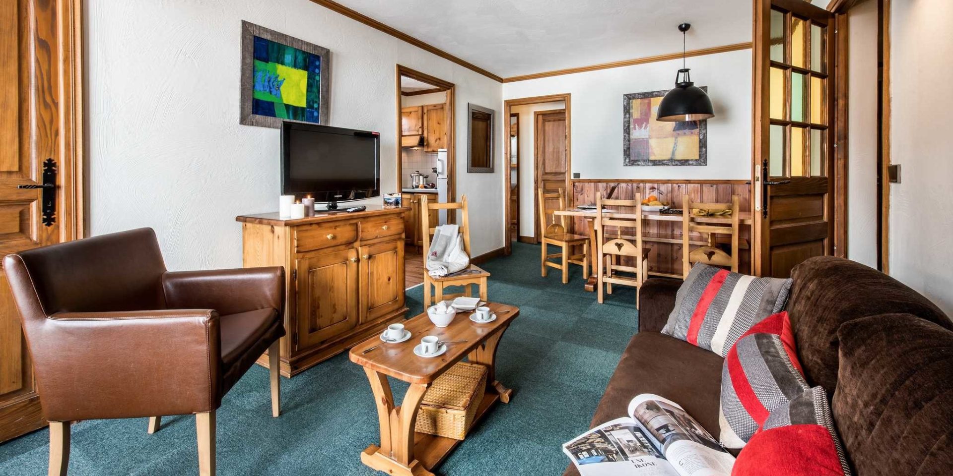 Residences Village Montana Val Thorens Les 3 Vallees Frankrijk wintersport skivakantie luxe living 4K6 bruie bank gestreepte kussens salontafel theekopjes bruine fauteuil TV schilderij eettafel houten stoelen hanglamp keuken hout