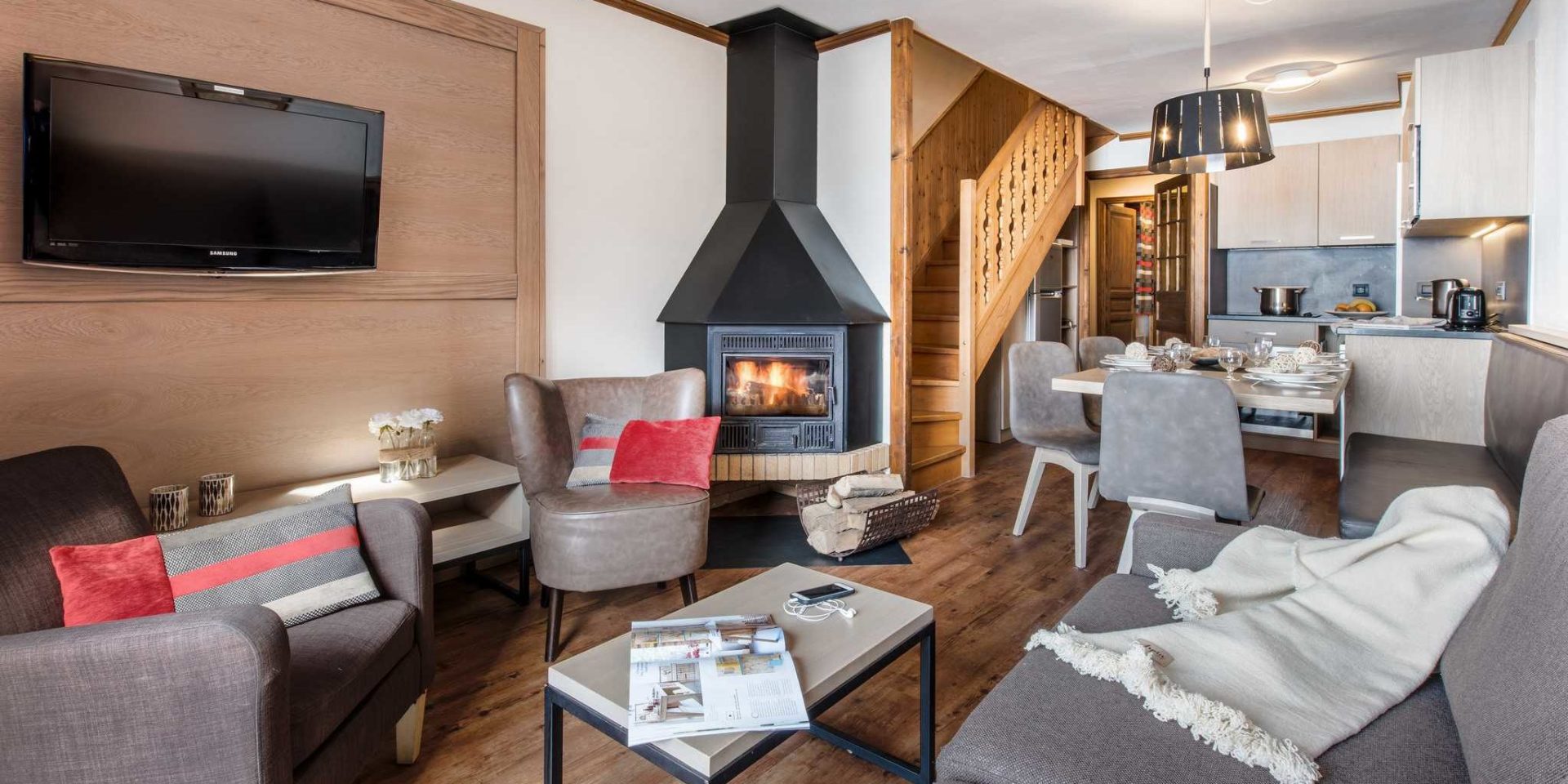 Residences Village Montana Val Thorens Les 3 Vallees Frankrijk wintersport skivakantie luxe living 4K6 66m2 grijze bank kleed fauteuils rode gestreepte kussens open haard hout salontafel boek gedekte tafel bankje stoelen trap keuken